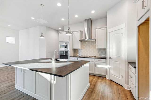 kitchen featuring a center island with sink, stainless steel appliances, white cabinetry, wall chimney range hood, and light hardwood / wood-style flooring