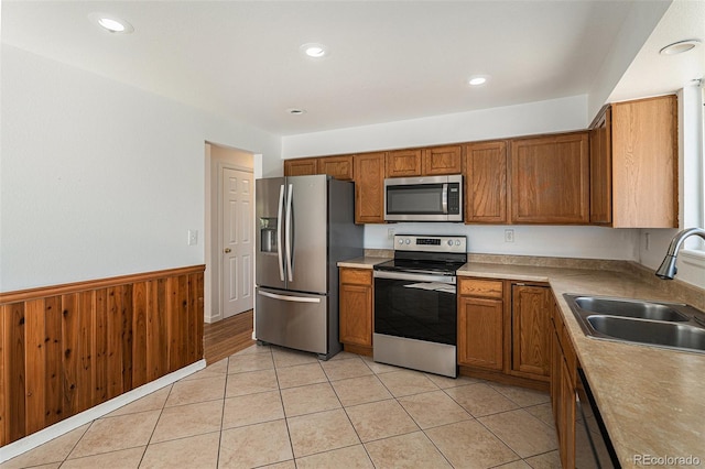 kitchen featuring light tile patterned floors, a wainscoted wall, a sink, appliances with stainless steel finishes, and brown cabinets