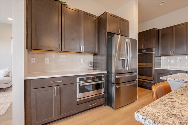kitchen with light stone countertops, dark brown cabinets, backsplash, light wood-style floors, and appliances with stainless steel finishes