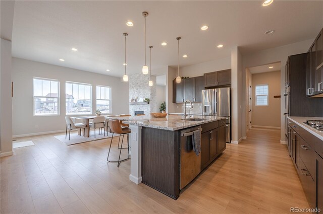 kitchen with a sink, appliances with stainless steel finishes, an island with sink, dark brown cabinetry, and light wood-style floors