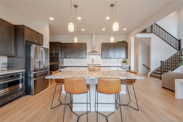 kitchen featuring stainless steel appliances, wall chimney exhaust hood, dark brown cabinets, a breakfast bar area, and light wood-style floors