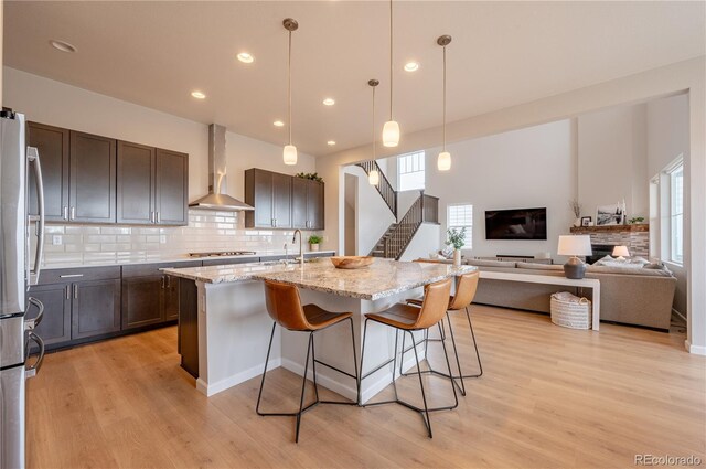 kitchen with light stone countertops, wall chimney exhaust hood, stainless steel appliances, a kitchen island with sink, and light wood-type flooring