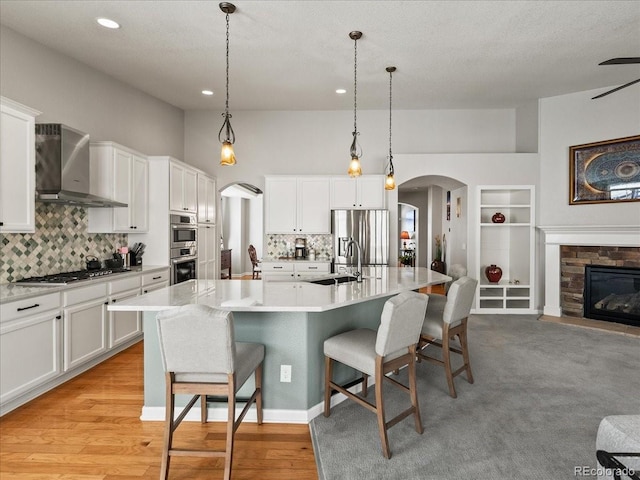 kitchen with arched walkways, stainless steel appliances, white cabinetry, wall chimney range hood, and a kitchen breakfast bar