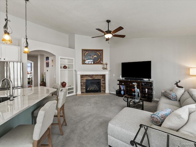 carpeted living area with baseboards, arched walkways, a ceiling fan, a stone fireplace, and built in shelves