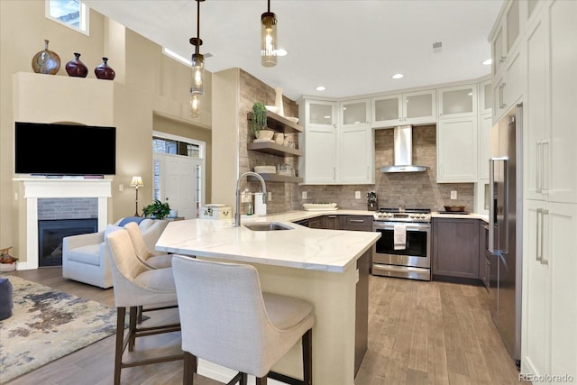 kitchen featuring a sink, stainless steel appliances, a peninsula, wall chimney exhaust hood, and light stone countertops