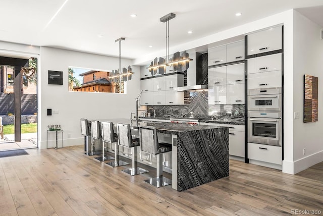 kitchen featuring an island with sink, wall chimney exhaust hood, white cabinetry, light wood finished floors, and baseboards
