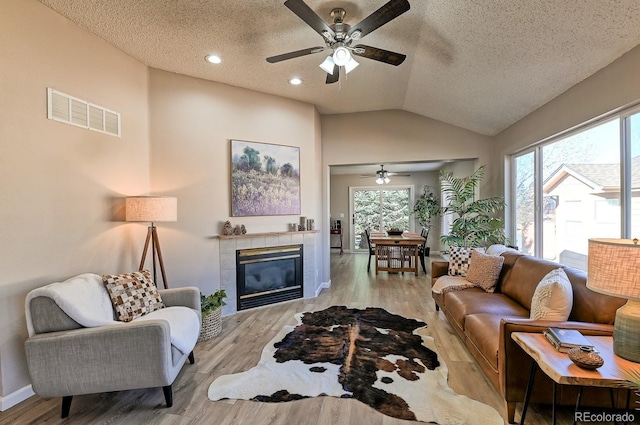 living room with lofted ceiling, ceiling fan, a textured ceiling, light hardwood / wood-style floors, and a tiled fireplace