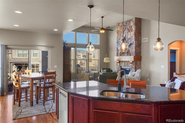 kitchen with sink, light hardwood / wood-style flooring, dark stone countertops, a center island with sink, and a stone fireplace