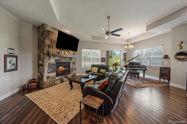 living room featuring ceiling fan with notable chandelier, a stone fireplace, a tray ceiling, and dark hardwood / wood-style flooring