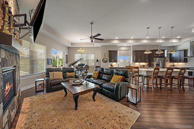 living room with baseboards, dark wood finished floors, a glass covered fireplace, a tray ceiling, and ceiling fan with notable chandelier