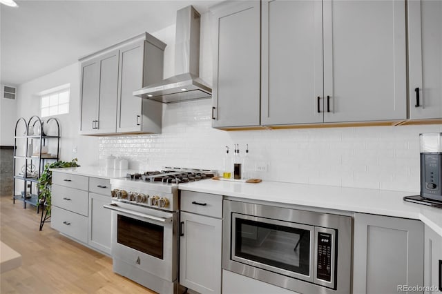 kitchen with gray cabinetry, appliances with stainless steel finishes, light wood-type flooring, and wall chimney exhaust hood