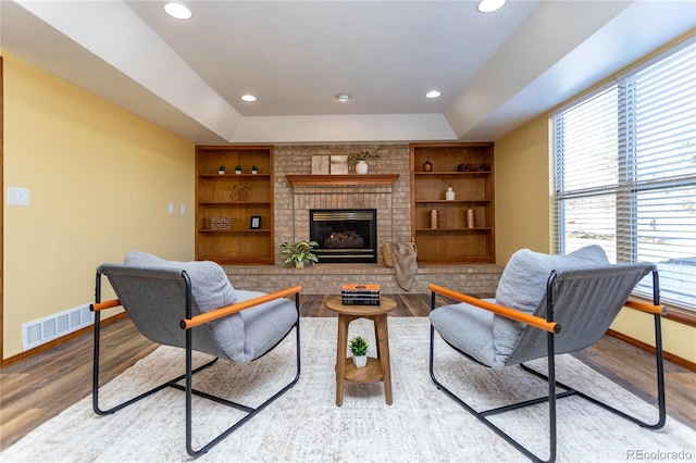 living room featuring a brick fireplace, built in shelves, a raised ceiling, and light wood-type flooring