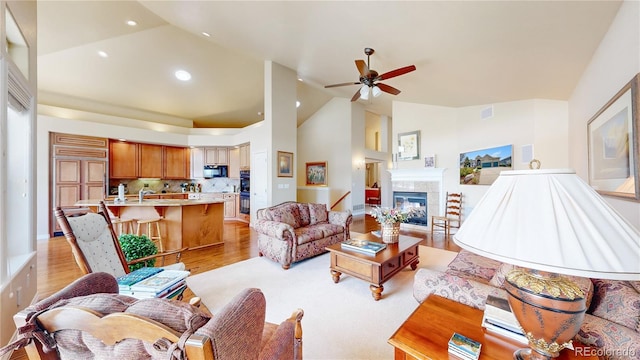 living room with a ceiling fan, a tiled fireplace, light wood-type flooring, high vaulted ceiling, and recessed lighting
