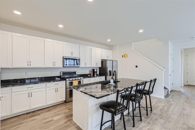 kitchen featuring stainless steel appliances, dark stone countertops, a center island with sink, and light wood-type flooring