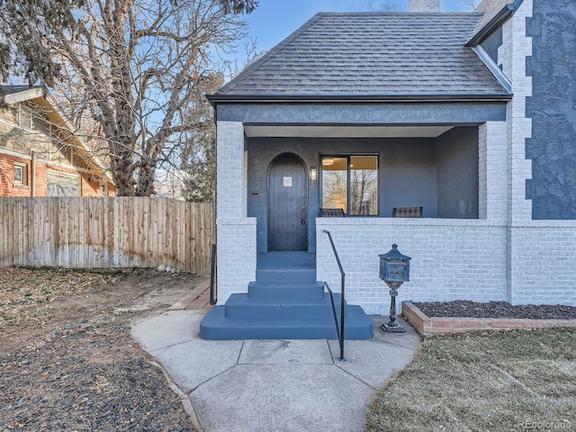 entrance to property featuring brick siding, roof with shingles, and fence