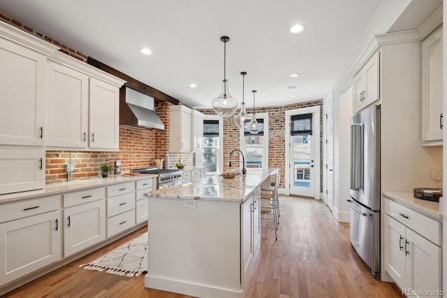 kitchen featuring high quality appliances, white cabinets, wall chimney exhaust hood, light wood finished floors, and an island with sink