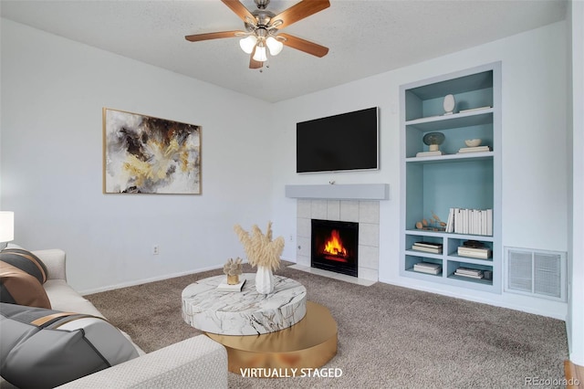 carpeted living room with ceiling fan, a tile fireplace, built in features, and a textured ceiling