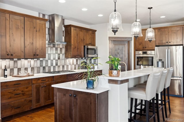 kitchen with wall chimney exhaust hood, decorative backsplash, hanging light fixtures, a center island with sink, and appliances with stainless steel finishes