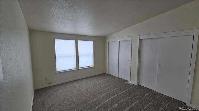 unfurnished bedroom with two closets, lofted ceiling, dark colored carpet, a textured wall, and a textured ceiling