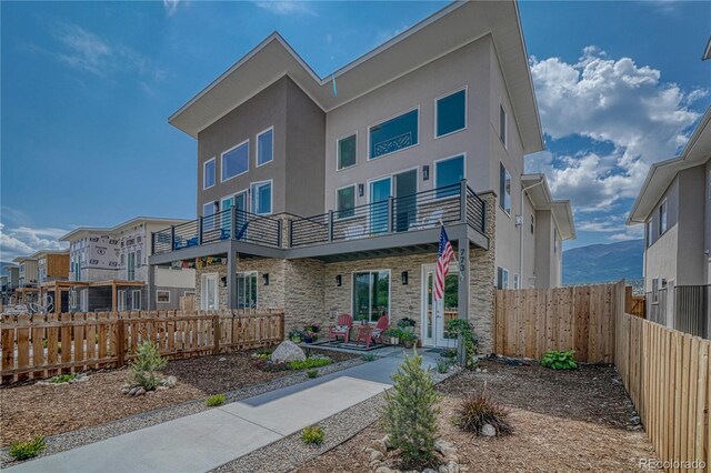 rear view of property with stone siding, fence private yard, a balcony, and stucco siding