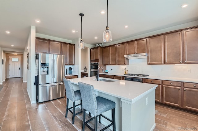 kitchen with an island with sink, under cabinet range hood, a sink, stainless steel appliances, and light wood finished floors