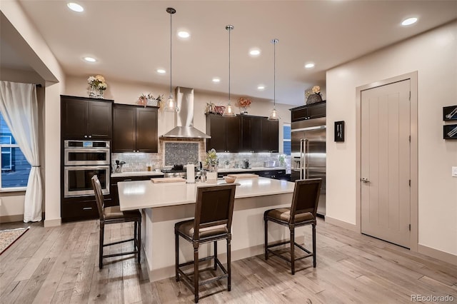 kitchen with a breakfast bar area, light countertops, backsplash, appliances with stainless steel finishes, and wall chimney range hood