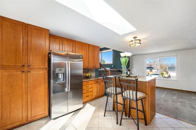 kitchen with light stone counters, a skylight, light colored carpet, brown cabinetry, and stainless steel fridge with ice dispenser