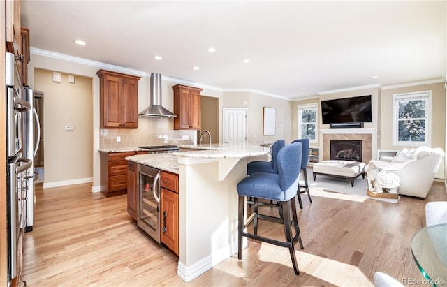 kitchen with wall chimney range hood, a breakfast bar, light stone counters, wine cooler, and a center island with sink