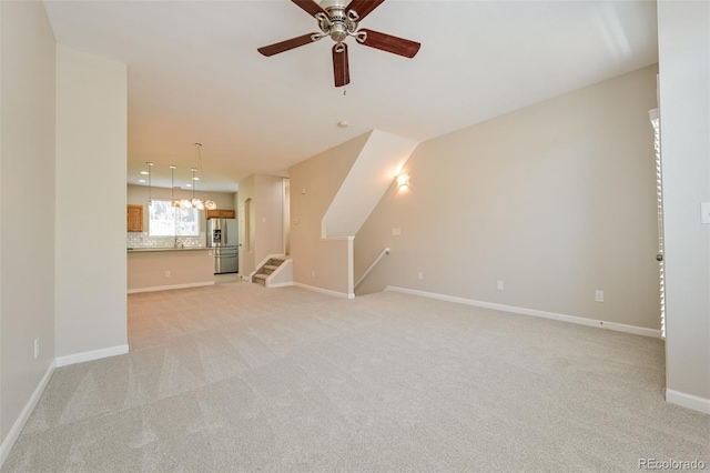 unfurnished living room with ceiling fan with notable chandelier, light colored carpet, and baseboards