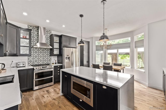 1588 W 111th Ave, Northglenn, CO 80234 - kitchen featuring appliances with stainless steel finishes, wall chimney exhaust hood, a center island, dark cabinetry, and light wood-type flooring