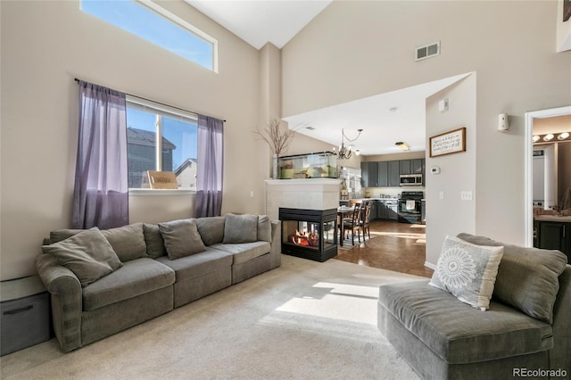 carpeted living room featuring a multi sided fireplace, a healthy amount of sunlight, a notable chandelier, and a towering ceiling