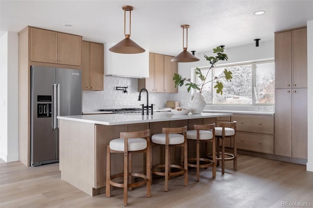 kitchen featuring high quality fridge, a kitchen island with sink, hanging light fixtures, light countertops, and backsplash