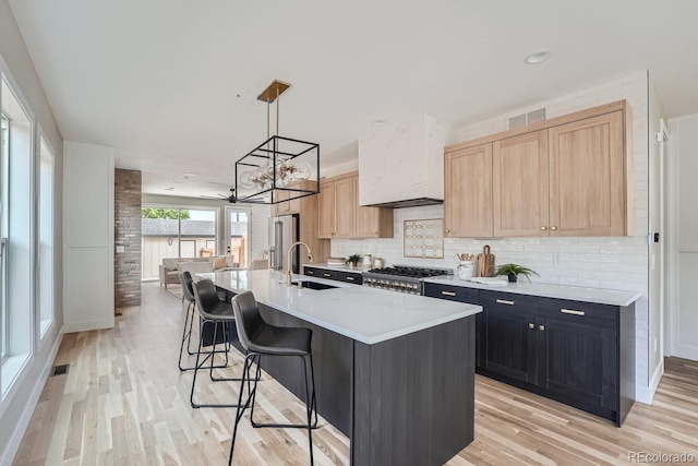 kitchen with light brown cabinetry, hanging light fixtures, sink, stove, and an island with sink