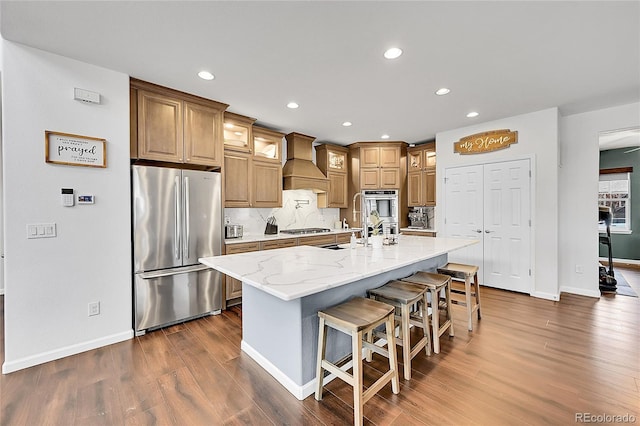 kitchen with dark wood finished floors, custom exhaust hood, stainless steel appliances, tasteful backsplash, and an island with sink