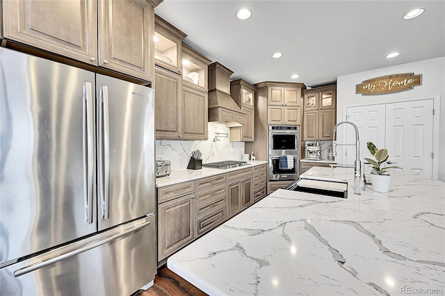 kitchen featuring light stone counters, custom exhaust hood, decorative backsplash, appliances with stainless steel finishes, and glass insert cabinets