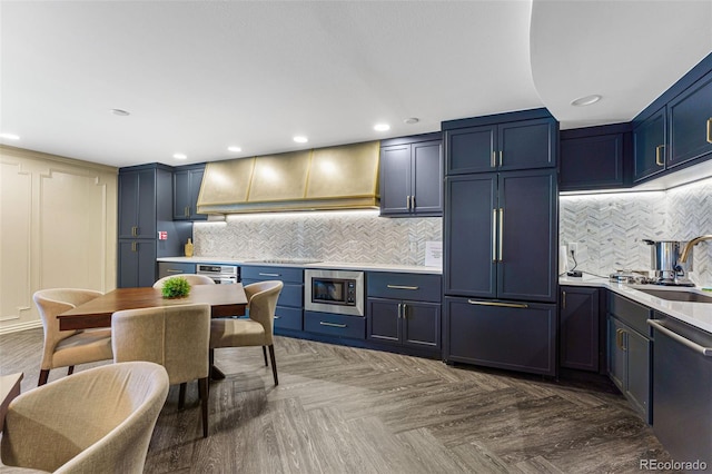 kitchen featuring built in appliances, custom exhaust hood, light countertops, blue cabinetry, and a sink