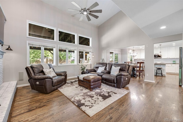 living area featuring ceiling fan, a high ceiling, wood finished floors, baseboards, and a brick fireplace
