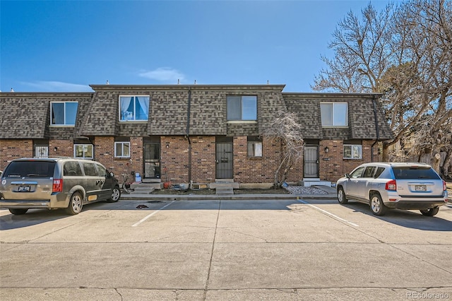 view of front facade with uncovered parking, mansard roof, a shingled roof, and brick siding