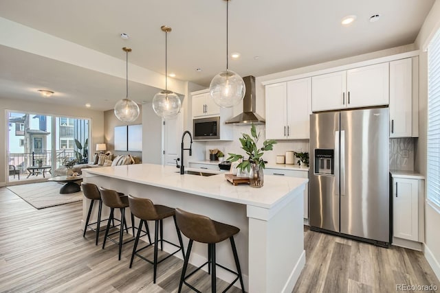 kitchen with wall chimney exhaust hood, a center island with sink, built in microwave, stainless steel refrigerator with ice dispenser, and white cabinetry