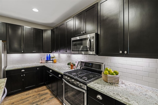 kitchen with light wood-style flooring, stainless steel appliances, dark cabinetry, light stone countertops, and tasteful backsplash