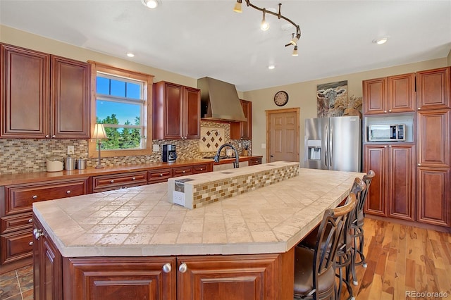 kitchen featuring a center island with sink, wall chimney exhaust hood, stainless steel appliances, and light hardwood / wood-style flooring