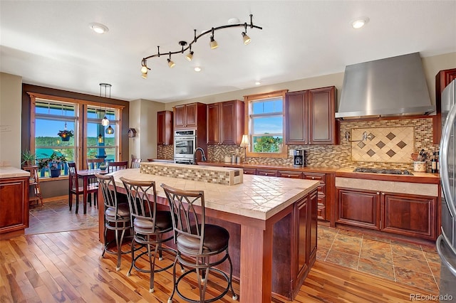 kitchen with a kitchen island, wall chimney exhaust hood, light hardwood / wood-style flooring, appliances with stainless steel finishes, and decorative backsplash