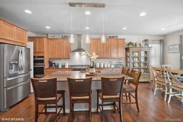 kitchen with dark wood-type flooring, a center island with sink, hanging light fixtures, wall chimney exhaust hood, and appliances with stainless steel finishes