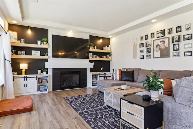 living room featuring a raised ceiling, a tiled fireplace, wood finished floors, built in shelves, and recessed lighting