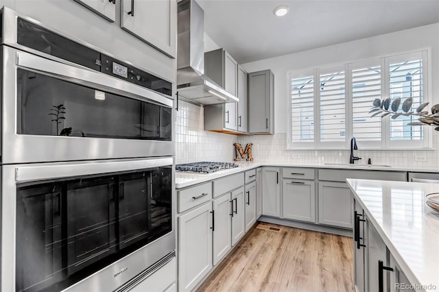 kitchen featuring sink, light wood-type flooring, gray cabinets, stainless steel appliances, and wall chimney range hood