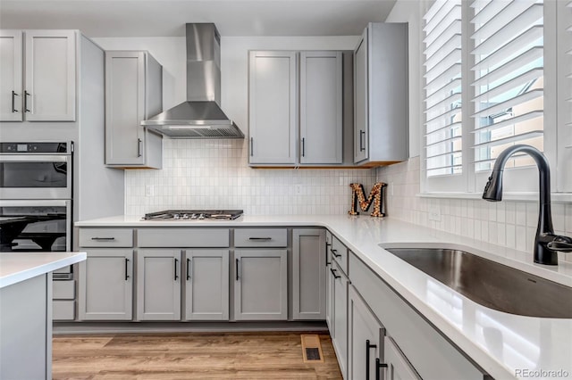 kitchen with gray cabinetry, wall chimney range hood, stainless steel appliances, and sink