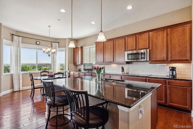 kitchen featuring an island with sink, stainless steel microwave, brown cabinets, hanging light fixtures, and backsplash