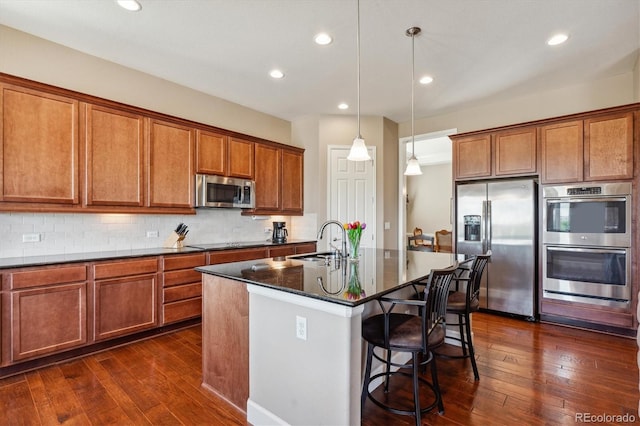 kitchen with dark hardwood / wood-style floors, stainless steel appliances, sink, pendant lighting, and a center island with sink