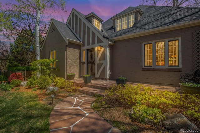 exterior entry at dusk with fence, stucco siding, roof with shingles, and brick siding