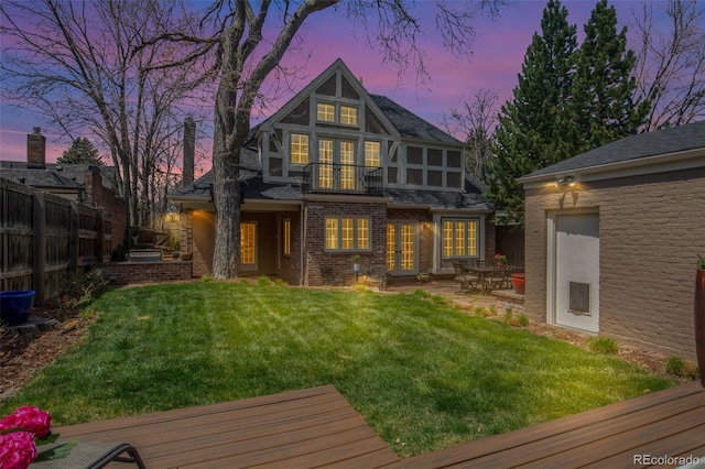 back of property at dusk featuring french doors, a lawn, brick siding, fence, and a balcony